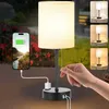 A hand is shown adjusting a bedside lamp that is charging a smartphone. The background shows a garden scene. Náhled obrázku produktu
