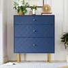 A navy blue dresser with gold legs and a decorative scale pattern on the drawers, adorned with a vase of flowers and books on top. Produktbild-Vorschau