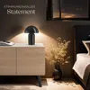 A bedside table with a lamp, book, and dried flowers next to a bed. Produktbild-Vorschau