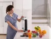 A woman is using a soda maker in a modern kitchen with fruits on the counter. Podgląd zdjęcia produktu