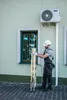 A worker is holding a folded ladder next to a building with an air conditioning unit. Anteprima dell’immagine prodotto