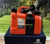 A Husqvarna combination fuel and oil canister sits on a dark blue box, with a blurred forest background. Voir les images du produit