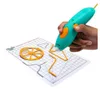 A hand is holding a teal and orange 3D printing pen, drawing on a white grid mat with pre-drawn outlines. There are already some yellow 3D printed objects on the mat. Podgląd zdjęcia produktu
