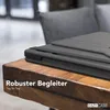 A black tablet case is shown on a wooden table, with a blurred background of a chair and room. Produktbild-Vorschau