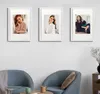 Three framed pictures of women are displayed on a white wall above two blue chairs. The chairs are arranged around a small wooden table with two mugs on it. A vase with dried flowers is visible on a small wooden stand to the right. Produktbild-Vorschau