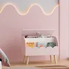 A pink toy chest with books and toys inside, placed in a child's room with a pink wall and a wavy light fixture. Náhled obrázku produktu