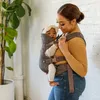 A woman is holding a baby in a carrier. There is a plant in the background. Produktbild-Vorschau