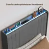 A close-up view of a modern upholstered headboard with a built-in shelf featuring integrated lighting, a digital clock, a smartphone, charging ports, and decorative items. Produktbild-Vorschau