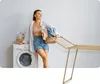 A woman is holding a basket of laundry next to a washing machine and a drying rack. Produktbild-Vorschau