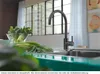 A black kitchen faucet is shown in a modern kitchen setting with fresh vegetables in the foreground and a large window in the background. Produktbild-Vorschau