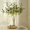 A vase of artificial olive branches with LED lights sits on a table next to a candle, with sheer curtains in the background. Produktbild-Vorschau