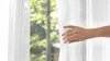 A hand is pulling back a white sheer curtain to reveal a blurred background of trees and foliage. Podgląd zdjęcia produktu