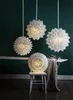 Four white paper star lanterns are displayed in a room with dark green walls. One lantern is on a chair in the foreground, and three are hanging from the ceiling in the background. Podgląd zdjęcia produktu