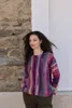 A woman stands in front of a stone wall and a white wall, wearing a colorful striped sweater. Produktbild-Vorschau