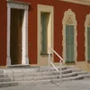 A stainless steel handrail is installed on a set of stone steps leading up to a red building with a wooden door and windows. Produktbild-Vorschau