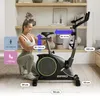 A woman is adjusting a stationary exercise bike in a room with shelves in the background. Dimensions are overlaid on the image. Produktbild-Vorschau