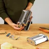 A person's hands are shown assembling a pasta maker on a wooden table, with other pasta maker parts and vegetables in the background. Anteprima dell’immagine prodotto