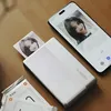 A Xiaomi portable photo printer is shown printing a photo of a young woman, with a smartphone displaying the same photo next to it. Several packs of ZINK photo paper are also visible. Anteprima dell’immagine prodotto