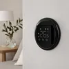 A black digital thermostat is mounted on a light-colored wall, with a bedside table and lamp visible in the blurred background. Náhled obrázku produktu