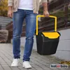 A person is walking on a paved path outdoors, carrying a black and yellow trash can with a yellow handle. Produktbild-Vorschau