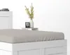 A close-up view of a white bed frame with a beige fitted sheet, next to a small white nightstand with a vase of yellow flowers and a framed picture. Anteprima dell’immagine prodotto