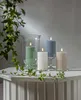 Four LED pillar candles of different colors and sizes are arranged on a white surface next to a glass hurricane lamp with a green candle inside. Green ivy vines drape over the scene. Podgląd zdjęcia produktu