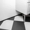 A close-up view of a black and white checkered floor with a white cabinet leg and a white wall. Náhled obrázku produktu