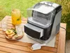 An ice maker sits on a wooden table outdoors, next to a pitcher of drink, glasses of ice, and fruit slices. Produktbild-Vorschau