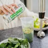 A person is pouring liquid from a can into a blender cup containing spinach and avocado. Produktbild-Vorschau