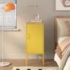 A yellow metal nightstand with a door and ventilation slots is placed next to a bed in a bedroom. A floor lamp with an orange shade is on the left, and a table lamp is on top of the nightstand. Podgląd zdjęcia produktu