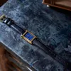 A rectangular gold-cased watch with a blue face and a black leather strap is laid out on a dark, textured surface. Produktbild-Vorschau