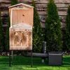 A wooden smoker oven is set up on a green lawn in front of a wooden fence and evergreen trees. Náhled obrázku produktu