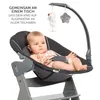 A baby sitting in a high chair attachment, with toys hanging above. Produktbild-Vorschau