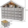 A white wooden advent calendar shaped like a house, decorated with miniature Christmas village elements and illuminated by warm LED lights. A single drawer is shown pulled out for scale. Voir les images du produit
