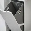 A close-up view of a white cabinet with a tilted open door, revealing a gray fabric laundry basket inside. Produktbild-Vorschau