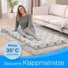 A woman is sitting on a folded mattress placed on a rug in a living room, next to a sofa. Produktbild-Vorschau