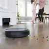 A robot vacuum cleaner is shown on a wooden floor with colorful candies scattered around it. In the background, two people are dancing, and a charging station for the robot is visible. Voir les images du produit