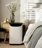 A white and black air purifier sits on a wooden nightstand next to a bed, with a vase of flowers on the nightstand. Anteprima dell’immagine prodotto