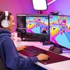 A woman wearing headphones is sitting at a desk and playing a video game on a computer. Produktbild-Vorschau