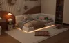 A bedroom scene with a corner bed, bedside table, lamp, and bookshelf. Podgląd zdjęcia produktu