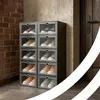 A stack of clear plastic shoe boxes filled with various pairs of sneakers, placed in a hallway next to a brick wall and a door. Podgląd zdjęcia produktu