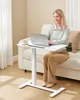 A woman is working on a laptop at a white adjustable height table next to a sofa. Produktbild-Vorschau