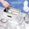 A hand is holding a necklace over an ultrasonic cleaning machine, with jewelry and accessories displayed around it. Produktbild-Vorschau