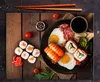 A top-down view of a breakfast meal with sushi, eggs, bacon, and soy sauce on a wooden table. Produktbild-Vorschau