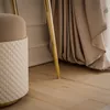 A close-up view of a quilted ottoman and the golden leg of a console table, with draped fabric and wooden flooring. Produktbild-Vorschau