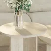 A close-up of a round travertine side table with a glass vase of flowers on it, placed next to a sofa. Produktbild-Vorschau