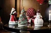 Christmas-themed decorative figurines including Santa Claus, a Christmas tree, and a snowman, arranged on a tray with a candle and a decorative orb. Produktbild-Vorschau