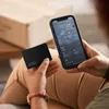 A person is holding a black card-shaped tracker and a smartphone displaying a map application. Produktbild-Vorschau