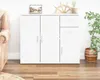 A white sideboard with doors and drawers, decorated with items on top, stands in a room with a couch and a plant. Produktbild-Vorschau