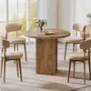 A round wooden dining table with four beige chairs in a bright, airy room with large windows. Produktbild-Vorschau